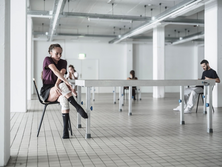 Eine Frau sitzt mit überschlagenen Beinen an einem Tisch. Im Hintergrund sitzen drei weitere Menschen im Raum verteilt an Tischen. Der Raum ist groß und weiß.