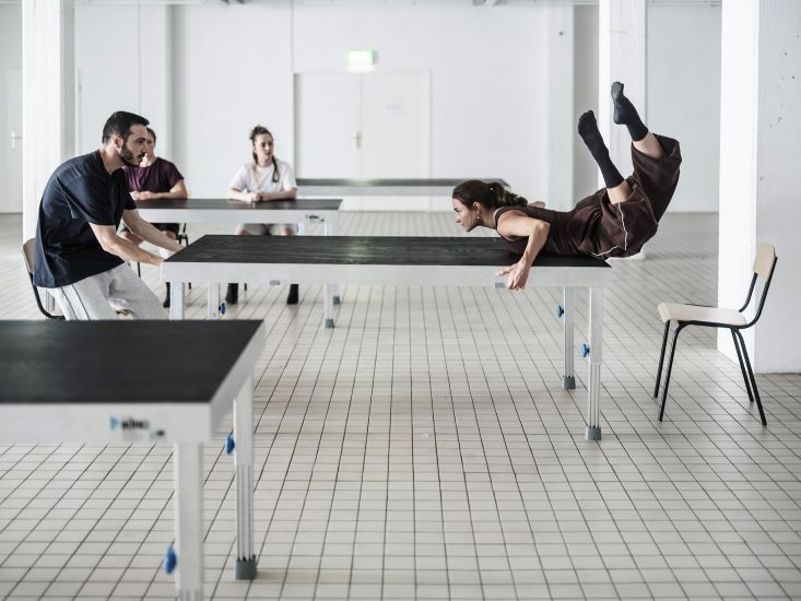 In einem weißen Raum zieht eine Person an einem Tisch. Auf der anderen Seite lehnt eine Frau über den Tisch. Ihre Beine sind in der Luft und sie schauen sich an. Im Hintergrund sitzen zwei Personen an einem Tisch und sehen sie an.