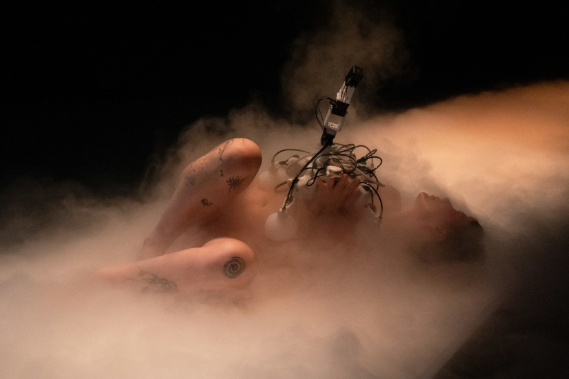 Eine nackte Frau liegt auf dem Boden. Um sie herum ist Nebel. Sie hält auf dem Oberkörper ein Gerät mit Kabeln.