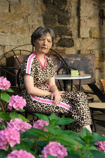 Eine Frau sitzt an einem Cafétisch. Sie trägt die Maske eines gealterten Gesichts. Im Vordergrund sind Blumen.