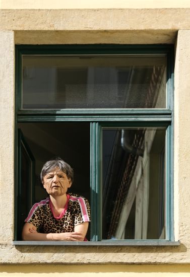 Eine Frau sieht aus dem Fenster. Sie trägt eine Maske eines gealterten Gesichts.