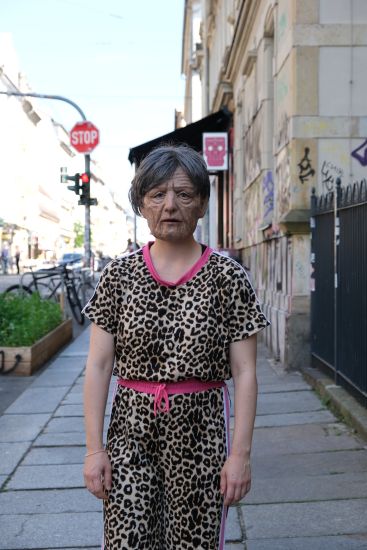 Eine Frau steht auf der Straße. Sie trägt Kleidung mit Leopardenmuster. Sie trägt eine Maske von einem gealterten Gesicht.