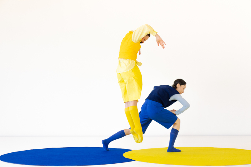 Auf dem Boden sind zwei große runde Kreise. Eine ist gelb, die andere blau. Eine Frau tritt von der blauen auf die gelbe Fläche. Sie trägt blaue Kleidung. Neben ihr springt ein Mann in die Luft. Er trägt gelbe Kleidung.