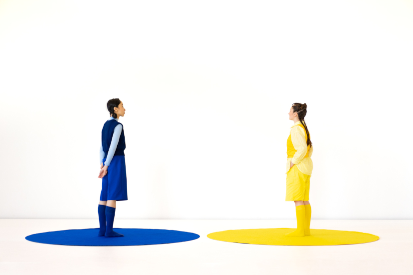 Zwei Frauen stehen in einem leeren Raum und blicken sich an. Die eine trägt blaue Kleidung, die andere gelbe Kleidung.