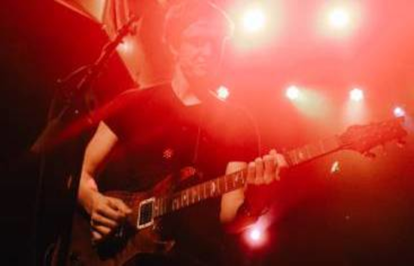 Foto von einem Mann, der E-Gitarre spielt. Er wird von hinten von roten Scheinwerfern angeleuchtet.