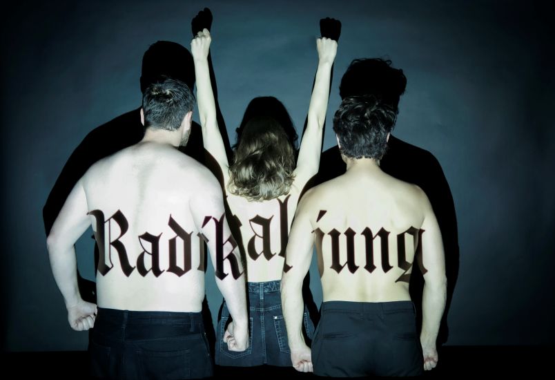 Foto von drei oberkörperfreien Menschen von hinten. Zwei Männer und eine Frau in der Mitte. Die Frau reckt ihre Fäuste in die Luft. Sie werden hell angeleuchtet. Auf ihren Rücken steht in Frakturschrift Radikal jung.